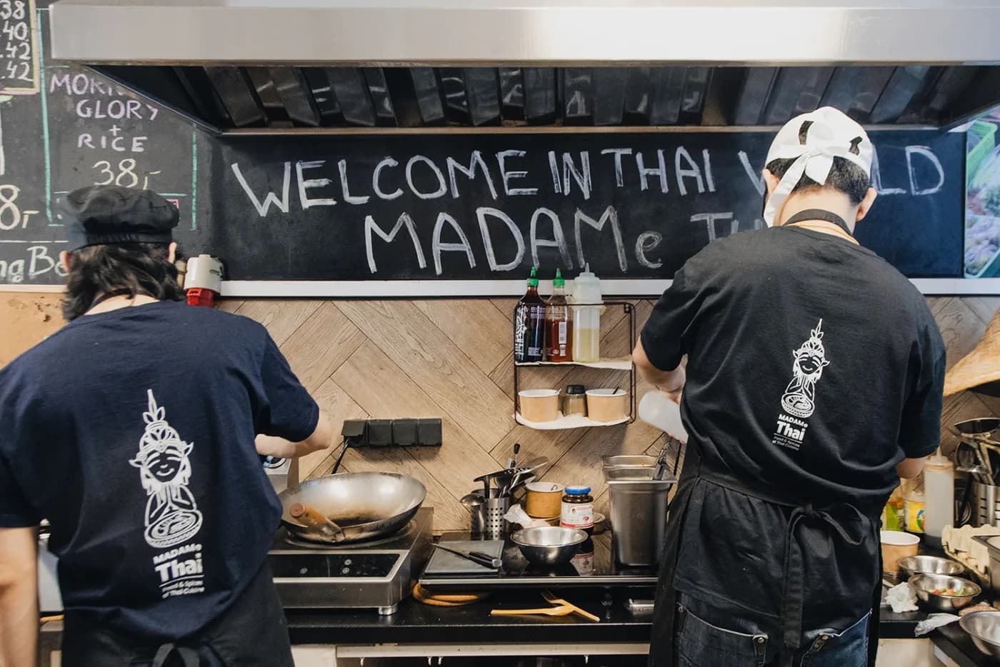 Restauracja tajska MADAMe Thai w Śródmieściu — otwarta kuchnia, kucharze przy wokach i tablica „Welcome in Thai World"